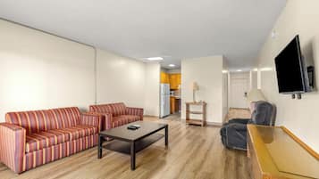 A living room with a sofa set, a coffee table, a TV mounted on the wall, and a kitchen in the background.