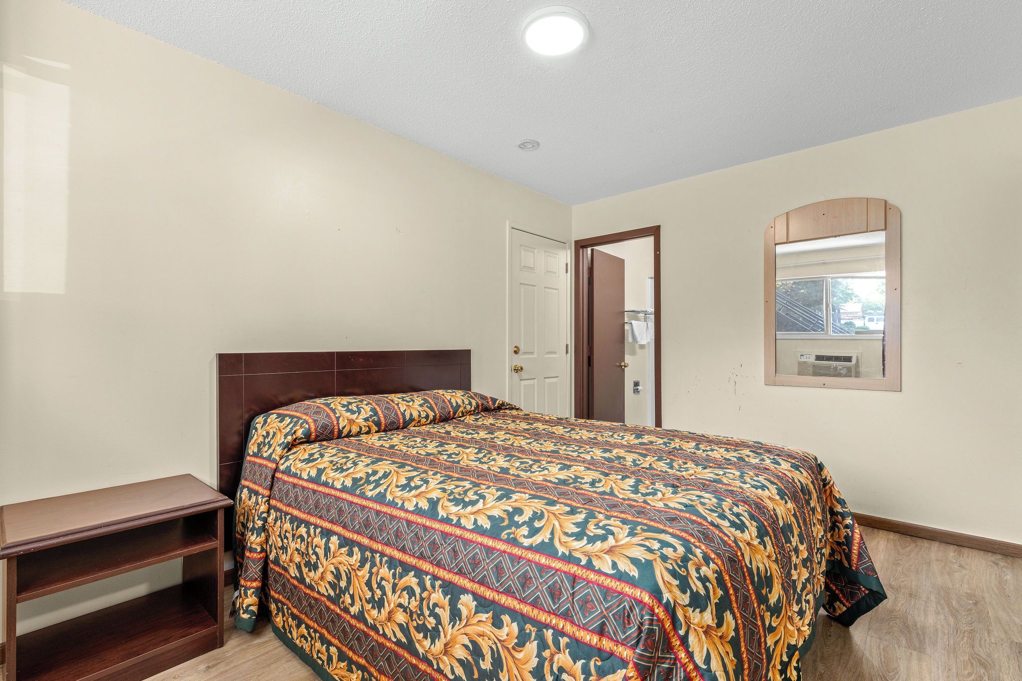 Quarto com cama, mesa de cabeceira de madeira, porta e espelho fixado na parede.