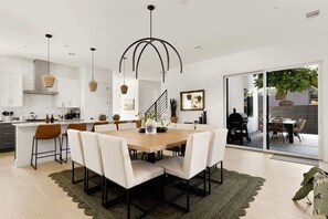 Eine moderne Küche mit weißen Schränken, ein Essbereich mit einem Holztisch und weißen Stühlen sowie eine große Schiebeglasdoor zum Außenbereich.