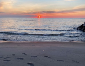 Ein Strand mit Fußspuren, die zum Wasser führen, ein Sonnenuntergang im Hintergrund und eine ruhige See.