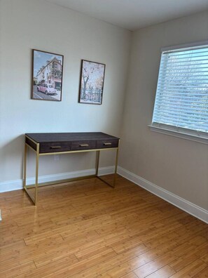 Ein Zimmer mit Holzboden, einem dunklen Schreibtisch mit goldfarbenen Füßen, zwei gerahmten Bildern an der Wand und einem Fenster mit Jalousien.