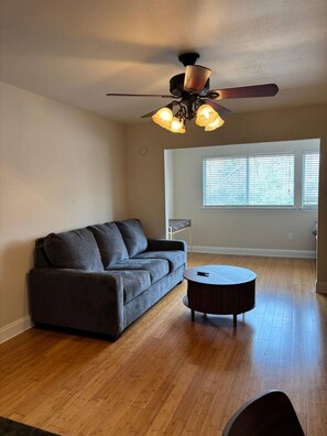 Ein Wohnzimmer mit grauem Sofa, rundem Couchtisch, Holzböden, Deckenventilator und einem Fenster mit Jalousien.