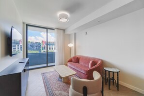 Moderni olohuone, jossa on sohva, sohvapöytä, televisio ja suuri ikkuna, josta avautuu näkymä rakennuksiin.