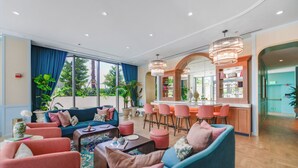 Een moderne zithoek met een blauwe bank, roze fauteuils, een bar met krukken en grote ramen voorzien van gordijnen.