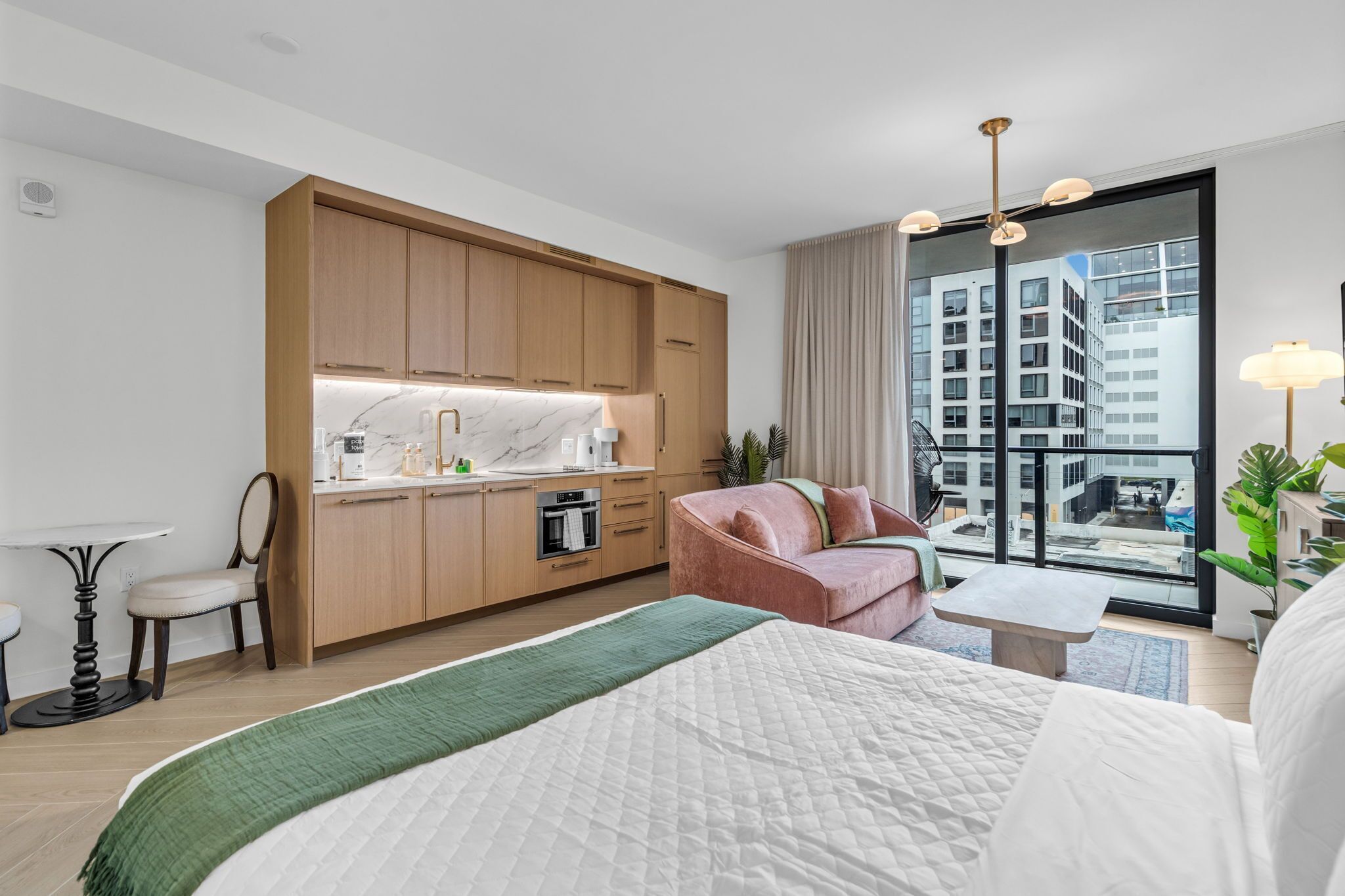A modern hotel room with a kitchenette, a bed, a seating area, and a large window with a city view.