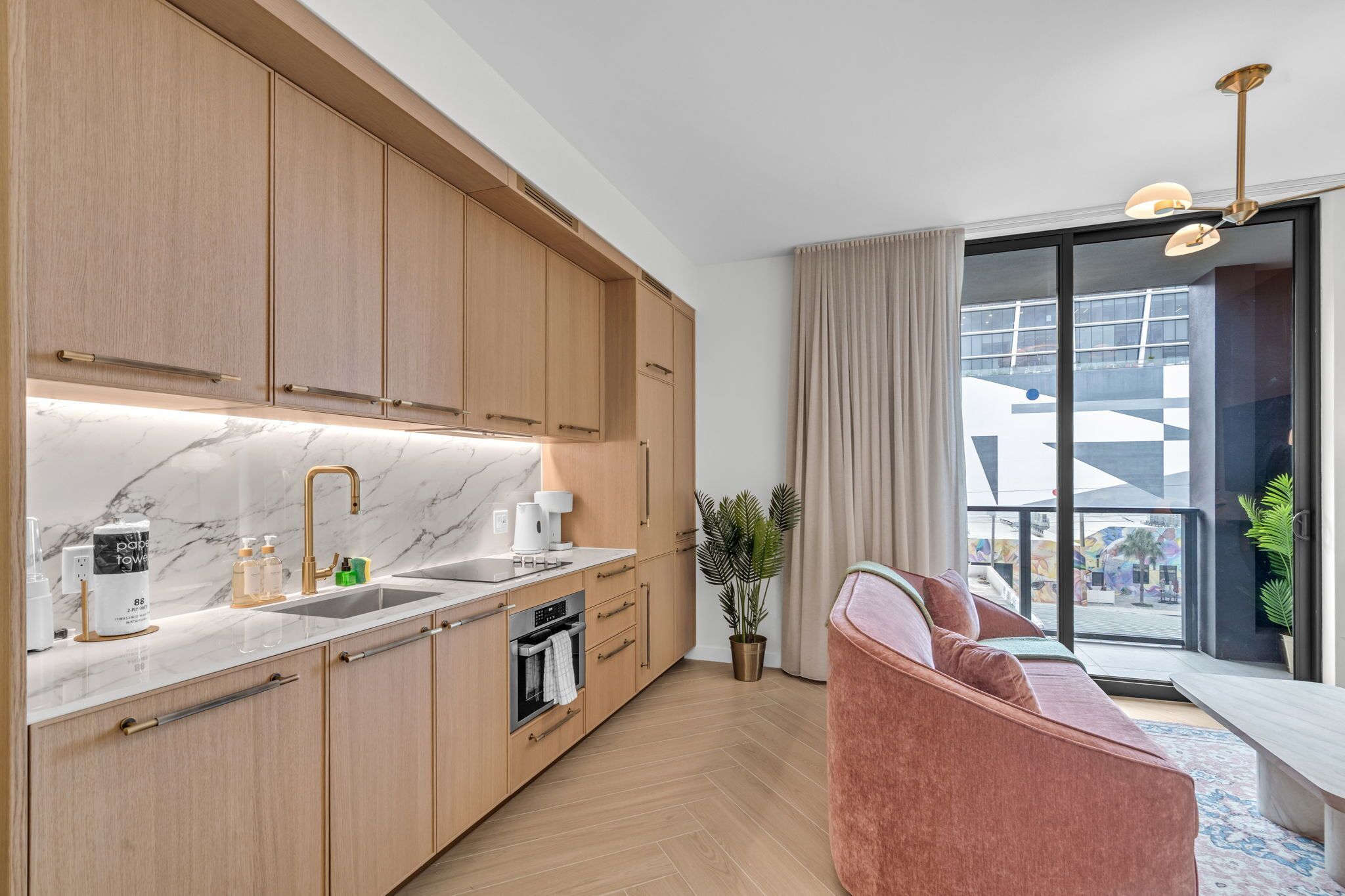 Eine moderne Küche mit Holzschränken, einer Marmorarbeitsplatte und einem eingebauten Ofen. Es gibt einen Essbereich mit einem rosa Sofa und Blick ins Freie.