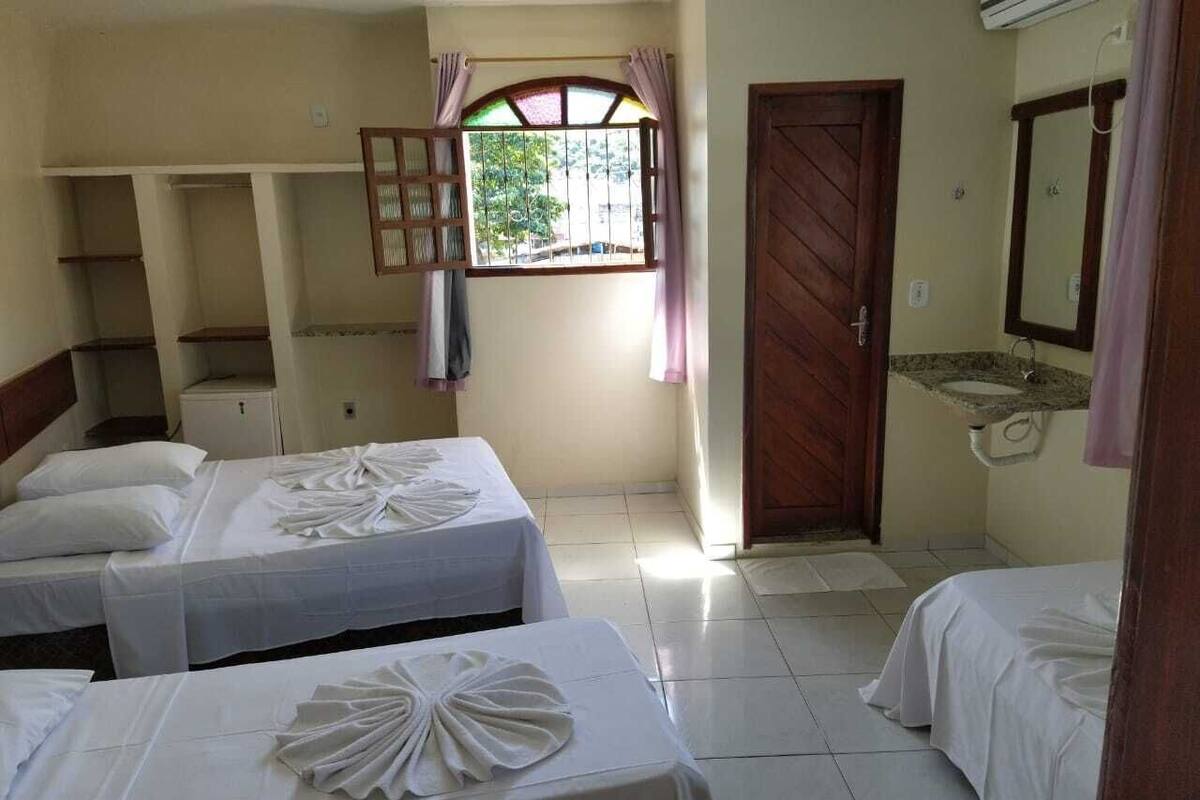 Habitación de hotel con tres camas, una ventana con cortinas, una puerta de madera, un lavamanos y un equipo de aire acondicionado.