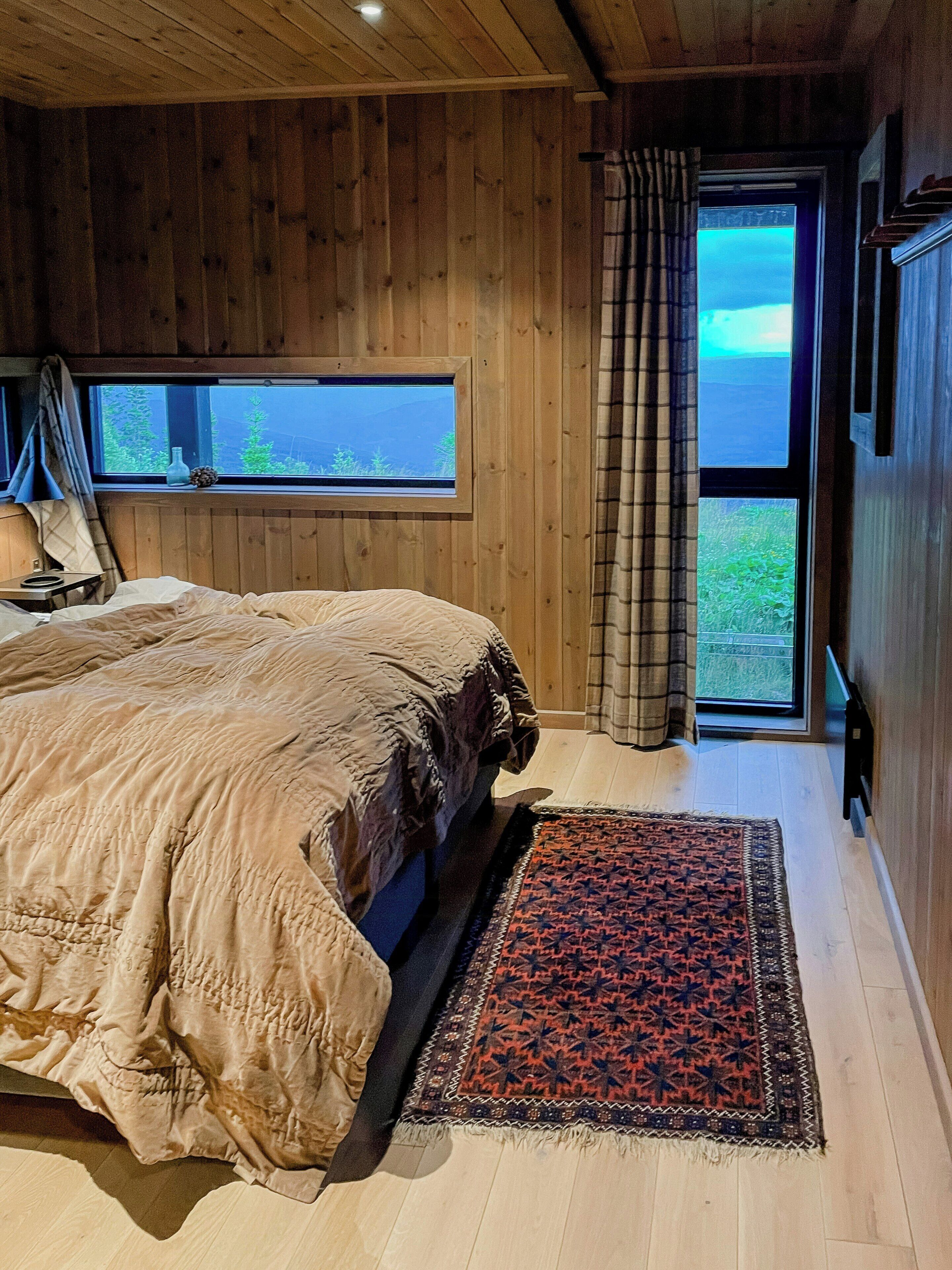 A wooden-paneled bedroom with a bed, a window with a view, and a door leading outside.
