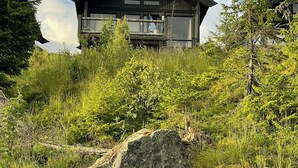 Een moderne berghut met balkon, omgeven door weelderige begroeiing en rotsen.