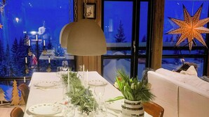 Een eettafel gedekt voor vier personen, met een witte tafeldoek, kaarsen en een vaas met groen.