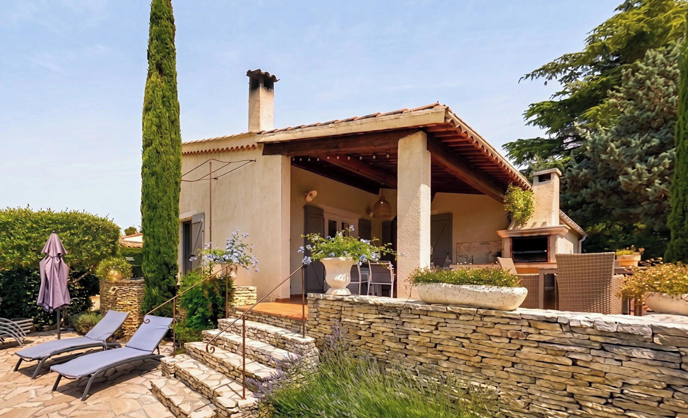A house with a stone wall, a chimney, a patio with lounge chairs, and a garden.