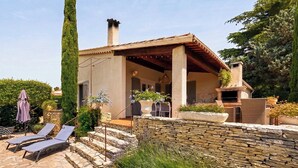Una casa con un muro in pietra, un camino, un patio con poltrone relax e un giardino.