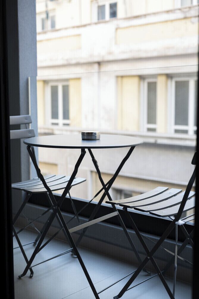A balcony with a round table and two chairs, overlooking a building with multiple windows.