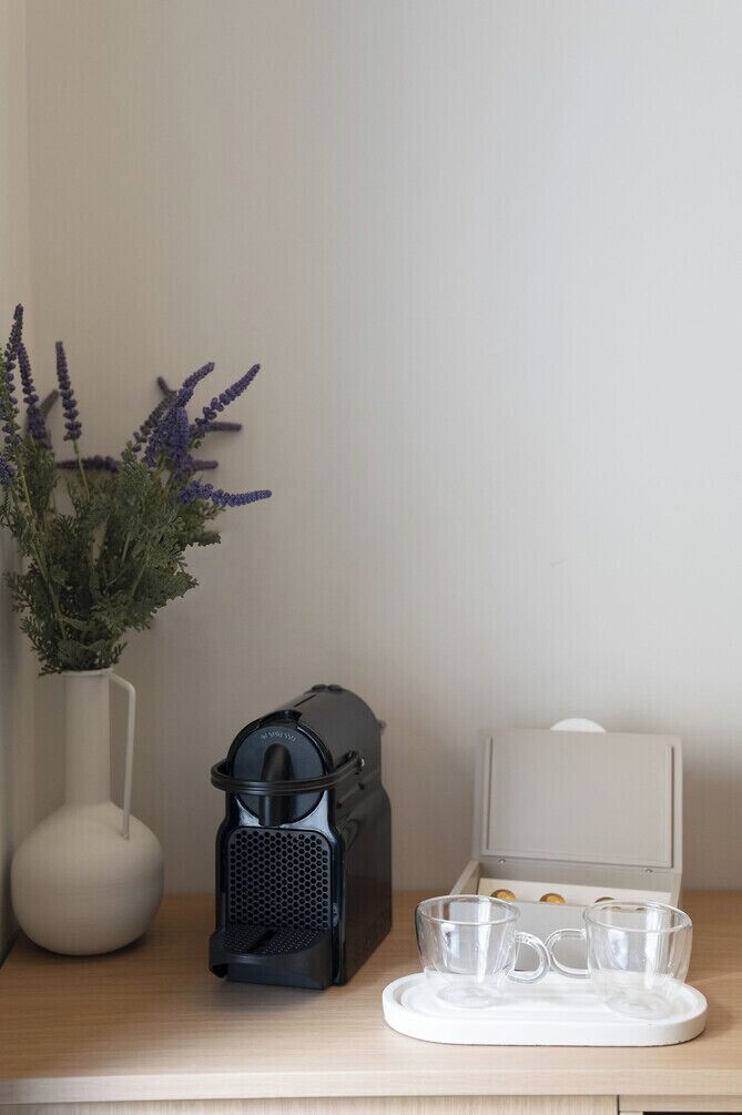A white vase with purple flowers, a black coffee maker, and a white tray with two glasses on a wooden surface.