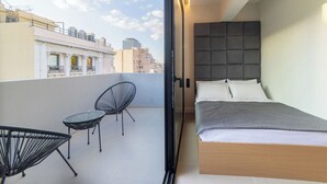 Ein modernes Schlafzimmer mit Bett, einem kleinen Balkon mit Stuhl und Blick auf die Stadt.