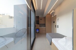 Ein modernes Zimmer mit Glaswand, einem Bett, einem Sessel, einem Fernseher und einer Küchenzeile.