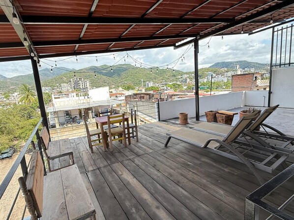 Une terrasse en bois avec une table et des chaises, un fauteuil de relaxation et une vue sur un paysage urbain avec des montagnes en arrière-plan.