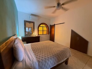 Un dormitorio con cama, cómoda, espejo, ventana con cortina, ventilador de techo y armario.