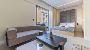 Een moderne hotelkamer met een bank, een bed, een tafel met een fles en glazen, en uitzicht op gebouwen door een raam.