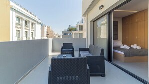 Een balkon met rieten meubilair, een bed met houten hoofdbord en uitzicht op gebouwen.
