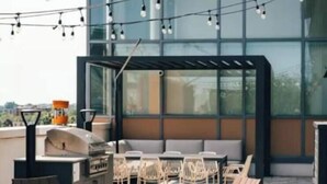 Uma área de estar externa com cadeiras e churrasqueira, com vista para um edifício moderno com grandes janelas.