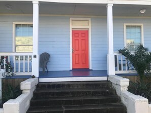 Uma casa azul com porta vermelha, colunas de varanda brancas e uma cadeira cinza no alpendre.