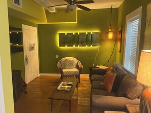 Ein Wohnzimmer mit Holzboden, einer Couch, einem Sessel, einem Couchtisch, einem an der Wand befestigten Regal mit Beleuchtung und einem Deckenventilator.