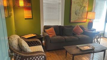A living room with a sofa, armchair, and coffee table. There is a painting on the wall and a lamp on a side table.