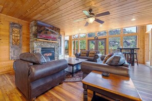 A cozy log cabin interior with a stone fireplace, leather sofas, and a dining area with a view of the surrounding forest.