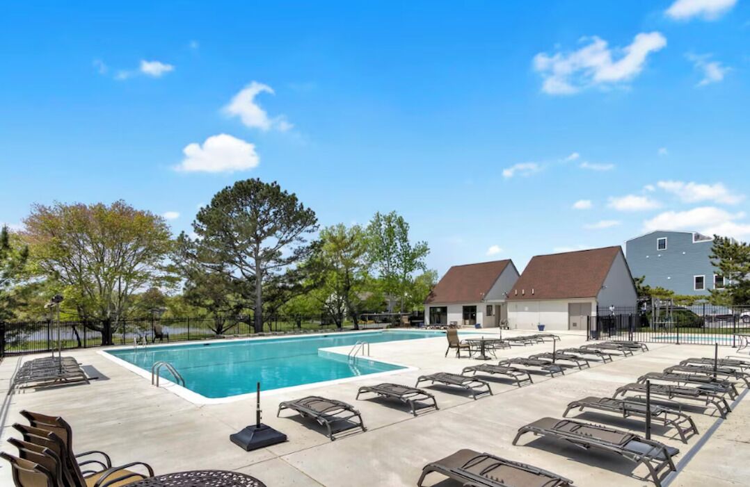 Une piscine avec des chaises longues, un bâtiment doté d’un toit rouge et des arbres en arrière-plan.