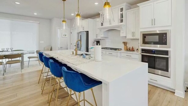 Cocina moderna con armarios blancos, una isla central con taburetes azules, electrodomésticos de acero inoxidable e iluminación colgante.