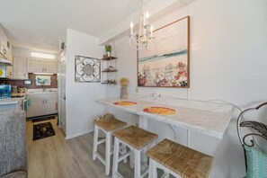 A kitchen with white cabinets, a marble countertop, and a chandelier. There are three stools and a painting on the wall. - Cozy 1 bedroom condo in enchanting Panama City Beach with AC (Panama City Beach)