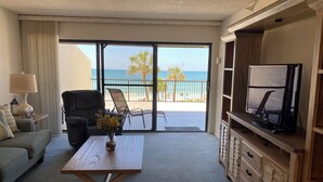 Un salon comprenant un canapé, un fauteuil, une table basse ornée de fleurs, un téléviseur posé sur un meuble en bois, et un balcon offrant une vue sur la plage et les palmiers.