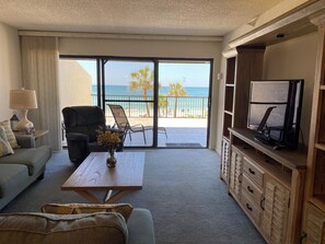 Ein Wohnzimmer mit Sofa, Sessel, Couchtisch mit Blumen, einem Fernseher auf einer Holzkommode und einem Balkon mit Blick auf den Strand und Palmen.