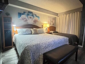 A hotel room with a large bed, a bench, a wardrobe, a nightstand with a lamp, and a wall with artwork. - Oceanfront Condo with Balcony, Pools & Walk to Boardwalk (Myrtle Beach)