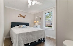 Ein Schlafzimmer mit Bett, Nachttisch, Lampe, Fenster und Deckenventilator.