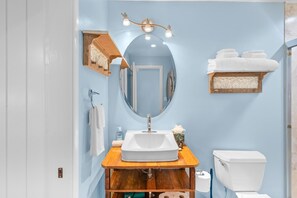 A bathroom with a wooden vanity, a white sink, a mirror with lights, a toilet, and a towel rack. - Kona Mansions#208 (Kailua-Kona)