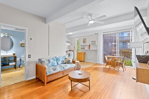 A spacious living area with a sofa, round table, and dining set. A ceiling fan and large windows are visible. - Kona Mansions#208 (Kailua-Kona)