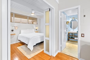 A bedroom with a bed, bedside lamps, a ceiling fan, and a view into the bathroom through an open door. - Kona Mansions#208 (Kailua-Kona)