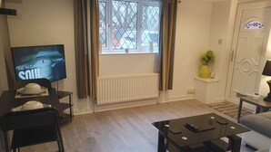 Una sala de estar con televisor, un radiador, una ventana con cortinas, un sofá, una mesa de centro y una puerta con un letrero.