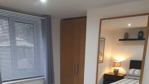Un dormitorio con cama, armario, mesita de noche con lámpara, espejo y ventana con persianas.