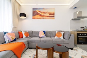 Un salon moderne avec un canapé d’angle gris, des coussins colorés, une petite table ronde, un tableau représentant un paysage désertique et un coin cuisine équipé d’une cuisinière et d’un four.