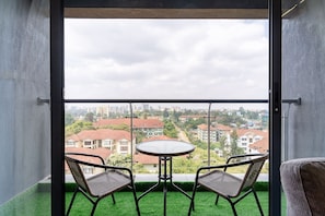 Un balcon avec une table ronde et deux chaises en osier, donnant sur un paysage urbain avec des bâtiments et de la verdure.