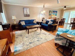 Ein Wohnzimmer mit blauem Sofa, einem Couchtisch, einem Esstisch mit Stühlen, einem Bild an der Wand und einem Deckenventilator.