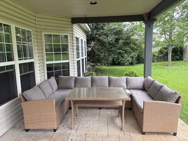 A covered patio with a sectional sofa and a table, surrounded by a house with large windows and a lawn.