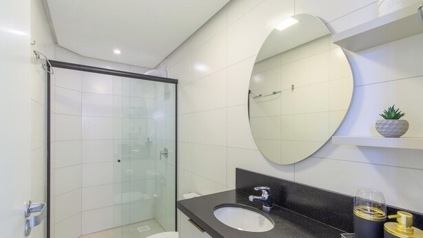 A modern bathroom with a glass shower enclosure, a round mirror, a black countertop, and a small shelf with a plant. - Apartment w/ Barbecue in Serra Gaucha Rad0501 (Canela)