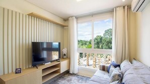 A modern living room with a flat-screen TV, a sofa, a window with a view of greenery, and a built-in shelf. - Apartment w/ Barbecue in Serra Gaucha Rad0501 (Canela)