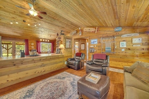 Ein Interieur einer Holzhütte mit Holzwänden, einem Deckenventilator, Ledersesseln, einem Sofa, einem Teppich und einer Theke mit rotem Vorhang.