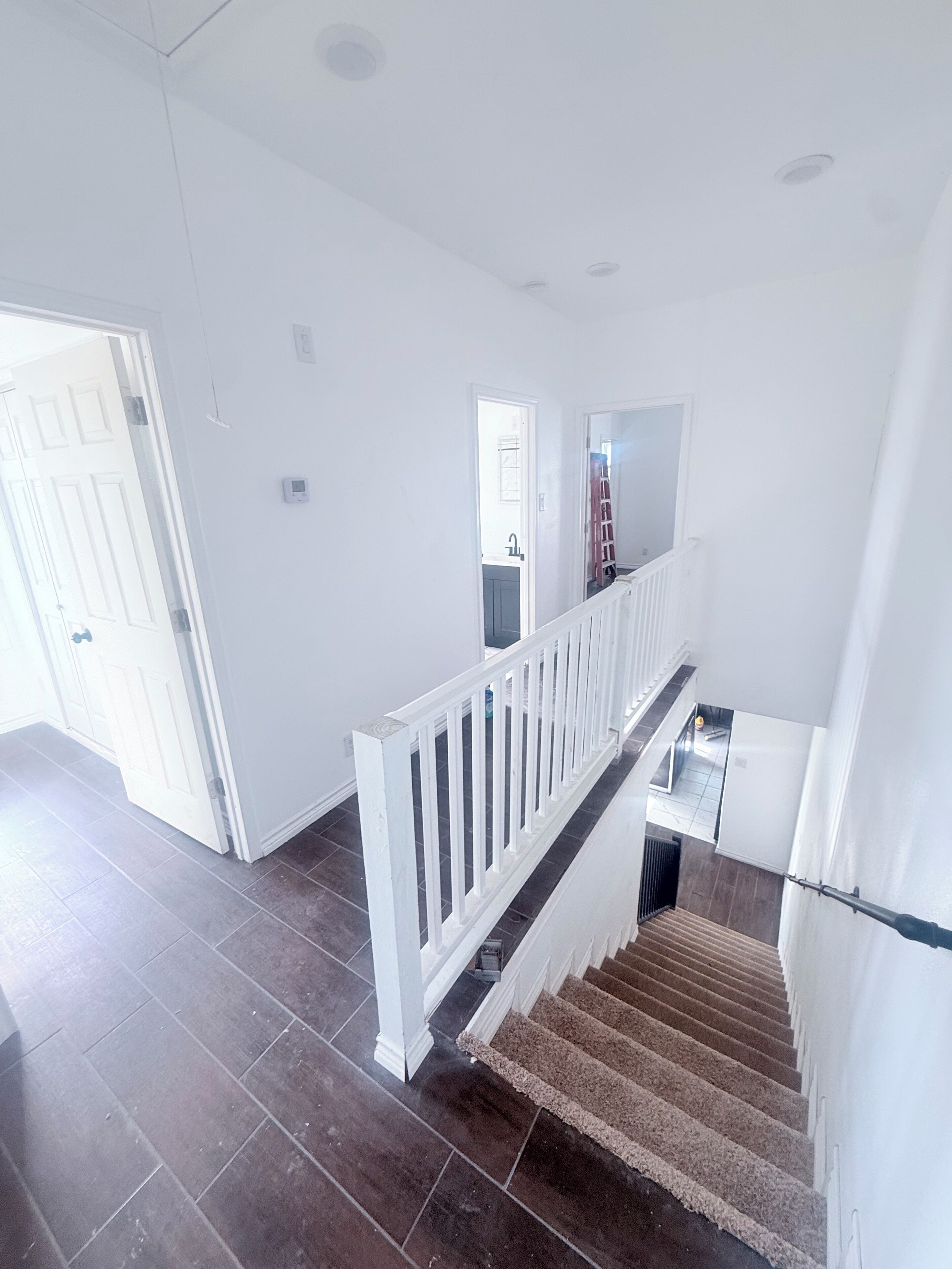 Un escalier avec une rampe blanche, un sol carrelé foncé et une vue sur une pièce avec une porte et une fenêtre.