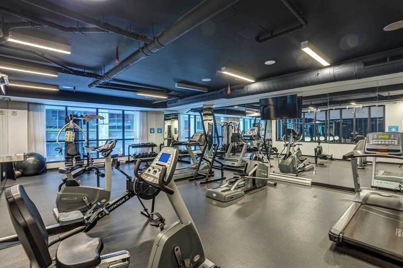 Un gimnasio con diversas máquinas de ejercicio, amplias ventanas y un espejo grande.